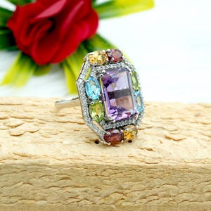 Könnte beinhalten: Ein silberner Ring mit einem großen violetten Amethyst-Edelstein, umgeben von kleineren Edelsteinen in verschiedenen Farben, darunter Blau, Grün, Gelb und Rot.
