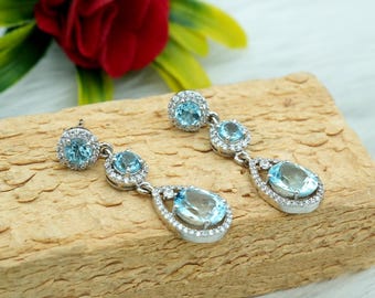 May include: A pair of silver dangle earrings with blue teardrop-shaped stones and smaller round stones. The earrings are on a wooden surface.