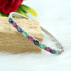May include: A silver bangle bracelet featuring a row of oval-shaped gemstones. The gemstones are blue, red, and green.