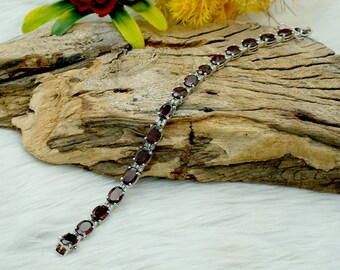 May include: A silver bracelet with a row of oval-shaped red gemstones. The bracelet is on a piece of driftwood.