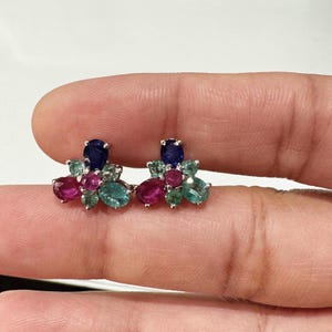 May include: A pair of stud earrings featuring a cluster of colorful gemstones. Each earring has a large oval-shaped, dark blue stone at the top, surrounded by smaller, oval-shaped pink and green stones. The earrings are set in a silver metal.