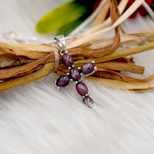 May include: A silver cross pendant featuring oval, deep purple gemstones. The cross is set against dried plant material and white, fluffy fabric. The pendant hangs from a thin silver chain.