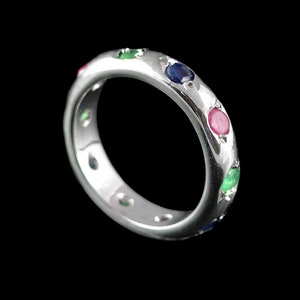 Può includere: Un anello a fascia d'argento con una fila di pietre preziose colorate. L'anello presenta smeraldi, zaffiri e rubini.