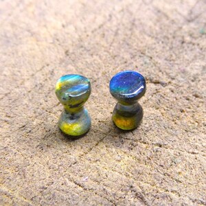 May include: Two iridescent blue and green glass plugs for stretched earlobes. The plugs are round and have a slight hourglass shape.