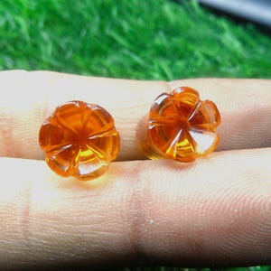 May include: Two orange, flower-shaped gemstones are displayed. The gemstones are held between two fingers. The gemstones have a faceted design, and the colour is a translucent orange. The background is blurred green.