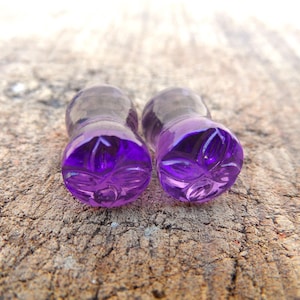 Może przedstawiać: Para kolczyków rozszerzających do uszu ze szkła w kolorze fioletowym z rzeźbionym wzorem kwiatowym.
