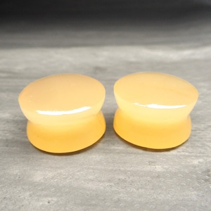 May include: A pair of smooth, translucent, light orange ear plugs with a concave center and flared edges. These circular body jewelry pieces have a glossy finish and a uniform, solid color throughout, designed for stretched ear piercings.