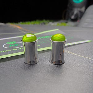 Könnte beinhalten: Zwei silberfarbene, zylindrische Objekte mit ausgestelltem Boden und einer hellgrünen, abgerundeten Oberseite. Die Objekte sind vor einem dunklen Hintergrund platziert, mit einer Visitenkarte im Hintergrund. Die Objekte scheinen dekorative oder funktionale Artikel zu sein.