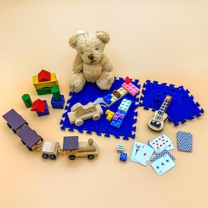 Può includere: Una collezione di giocattoli per bambini tra cui un orsacchiotto, un treno di legno, blocchi di costruzione, una chitarra giocattolo, carte da gioco e un cavallo a dondolo di legno. I giocattoli sono disposti su un tappetino da gioco blu e bianco.