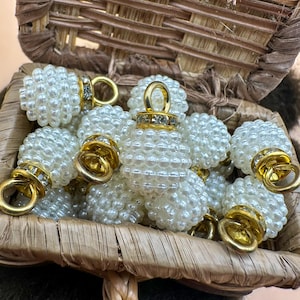 Peut inclure: Un assortiment de perles blanches en forme de perles avec des accents et des boucles dorées, nichées dans un panier tressé. Les perles ont un design décoratif, certaines sont ornées de strass transparents.