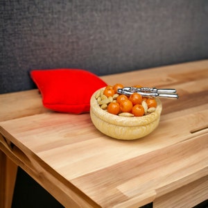 Op de afbeelding: Een houten kom gevuld met oranje en bruine snoepjes, een zilveren notenkraker en een rood kussen op een houten tafel.