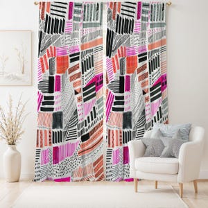 Op de afbeelding: Een paar gordijnen met patroon en een witte achtergrond. Het ontwerp toont een patchwork van geometrische vormen in zwart, grijs, roze en oranje. De gordijnen hangen voor een raam, met een witte stoel en kussens op de voorgrond.