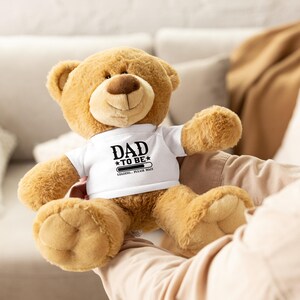 May include: A plush, light brown teddy bear wearing a white t-shirt with the text "DAD TO BE" and a loading bar graphic. The bear has a brown nose and is sitting with its arms outstretched.