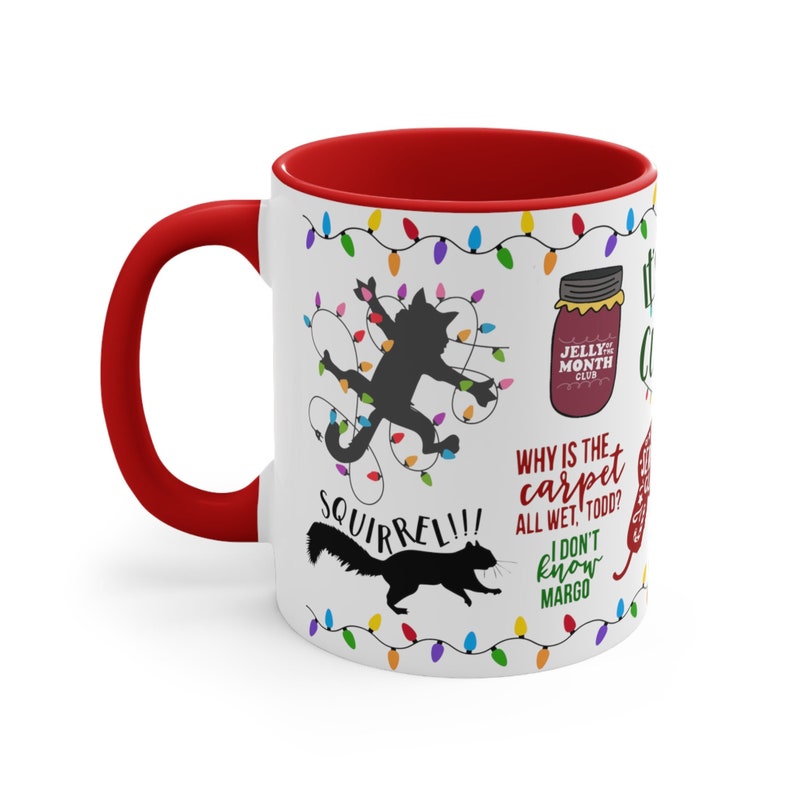 May include: White ceramic mug with a red interior and handle. The mug features various cartoon graphics and text, including a black cat tangled in Christmas lights, a squirrel, and the phrase "Squirrel!!!". Additional text includes "Jelly of the Month Club".