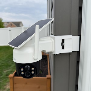May include: A white and black security camera with a solar panel attached, mounted on a gray wall. The camera has a black lens and is positioned above a wooden planter box. The sky is overcast.