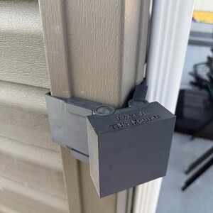 May include: A dark gray plastic mount attached to the corner of a beige vinyl siding wall. The mount features a hinged design with a square front face. The text "Made in USA" and "TNS Mounts" is embossed on the top surface of the mount.