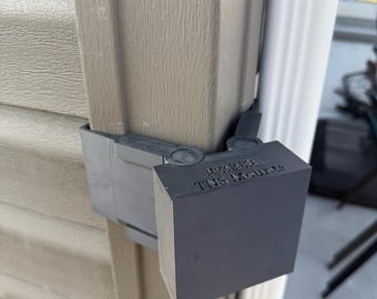 May include: A dark gray plastic mount attached to the corner of a beige vinyl siding wall. The mount features a hinged design with a square front face. The text "Made in USA" and "TNS Mounts" is embossed on the top surface of the mount.