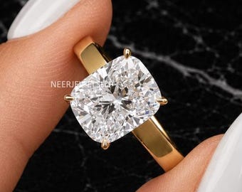 May include: A gold ring featuring a large, cushion-cut clear gemstone held by four prongs. The band is smooth and polished gold. The text NEERJEWELS is visible on the side of the setting. The design highlights the clarity and facets of the stone.