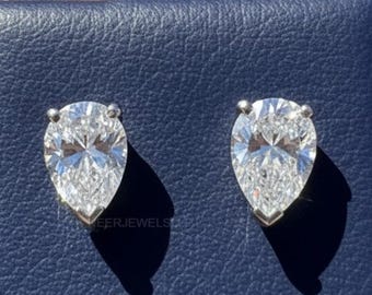 May include: A pair of pear-shaped diamond stud earrings. The diamonds are set in a silver metal and are displayed on a blue surface. The earrings are shown in a close-up shot.