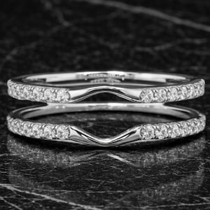 Puede incluir: Dos anillos plateados curvados, cada uno con una fila de pequeñas piedras transparentes. Los anillos están diseñados para encajar. El fondo es una superficie oscura y veteada, creando contraste con los anillos brillantes.