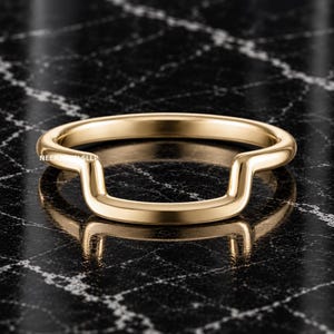 Puede incluir: Un anillo de oro pulido con una banda lisa y continua que presenta una distintiva hendidura o contorno en forma de U en un lado. La superficie metálica es reflectante y de color uniforme. El texto NEERJEWEL SLLP está impreso en letras blancas.