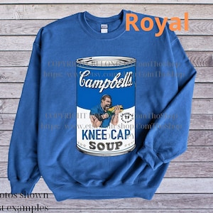 Op de afbeelding: Een koninklijk blauw sweatshirt met een afbeelding van een Campbell's soepblik met een man die een knieschijf vasthoudt en de tekst "Knee Cap Soup".