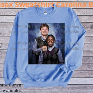 May include: Carolina Blue unisex sweatshirt with a printed image of two people wearing argyle vests. The sweatshirt has a crew neck and long sleeves. The text "Unisex Sweatshirt Carolina Blue" is at the top.