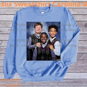 May include: Carolina blue unisex sweatshirt with a crew neck. The sweatshirt features a group photo of three people. The text "Unisex Sweatshirt Carolina Blue" is printed at the top of the image.