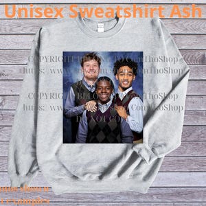 May include: A light gray unisex sweatshirt with a graphic of three people. The sweatshirt has the text "Unisex Sweatshirt Ash" at the top. The graphic is a portrait of three people wearing argyle vests and button-down shirts.