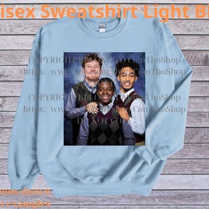 May include: Light blue unisex sweatshirt with a crew neck. The sweatshirt features a printed image of three people. The text "Unisex Sweatshirt Light Blue" is printed at the top. The background is a wooden surface.