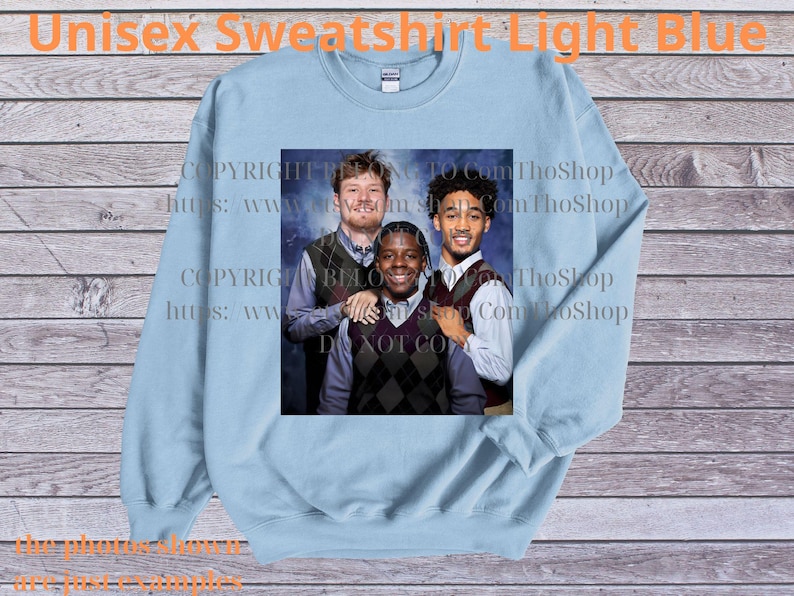 May include: Light blue unisex sweatshirt with a crew neck. The sweatshirt features a printed image of three people. The text "Unisex Sweatshirt Light Blue" is printed at the top. The background is a wooden surface.