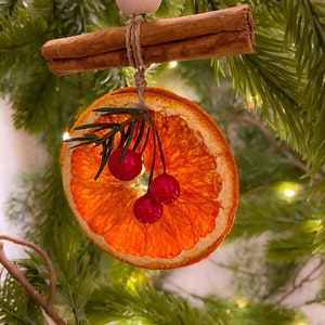 May include: A Christmas decoration comprising a dried orange slice, cinnamon sticks, and red berries, suspended by twine and a wooden bead. The ornament hangs against a backdrop of evergreen branches. The orange slice is approximately 7.6 cm in diameter.