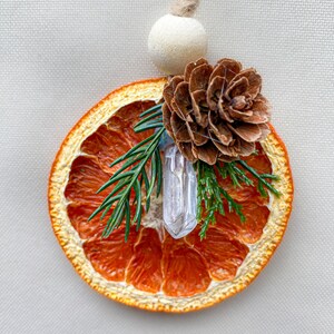 May include: A decorative ornament featuring a dried orange slice, a clear crystal, a pine cone, and evergreen sprigs. It hangs from a natural twine cord and a wooden bead. The orange slice is a vibrant orange colour.