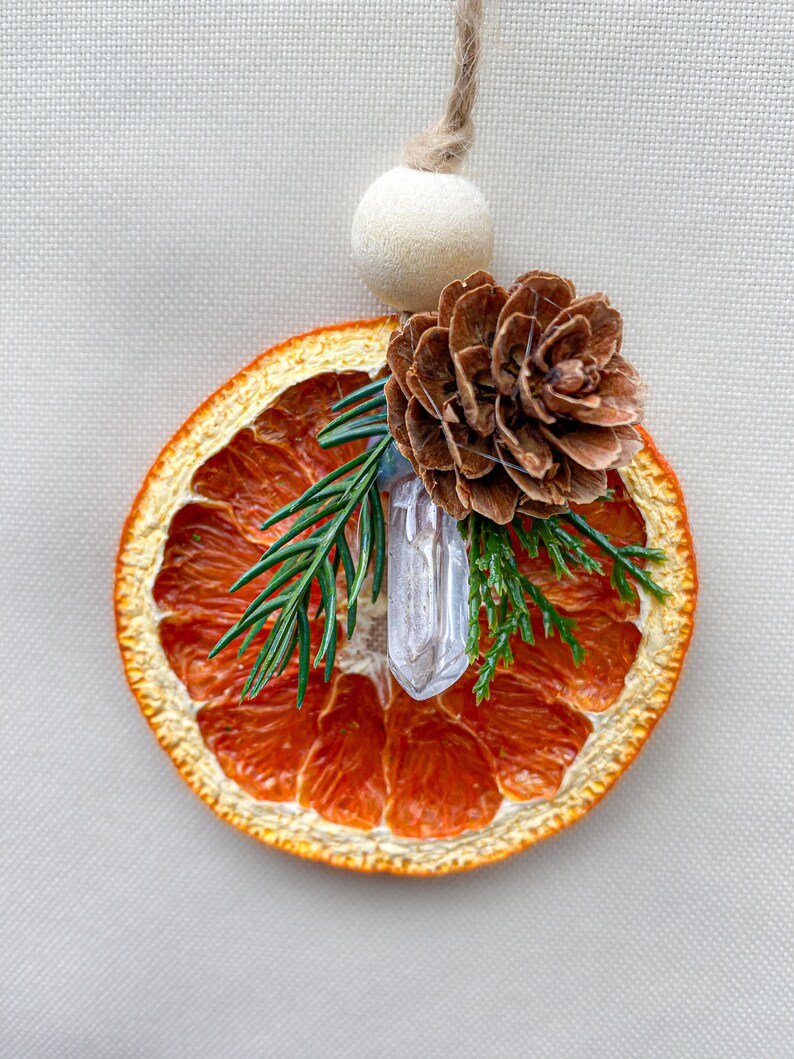 May include: A decorative ornament featuring a dried orange slice, a clear crystal, a pine cone, and evergreen sprigs. It hangs from a natural twine cord and a wooden bead. The orange slice is a vibrant orange colour.