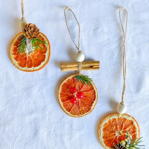 May include: Three festive Christmas ornaments crafted from dried orange slices. Each features natural decorations such as pine cones, cinnamon sticks, berries, and greenery. They are suspended by twine with wooden beads, offering a rustic, seasonal aesthetic.