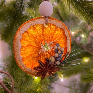 May include: A festive Christmas tree ornament featuring a dried orange slice, star anise, and berries. The orange slice is a bright orange colour and is translucent. It's suspended by twine and a wooden bead. The ornament hangs on a green Christmas tree branch.