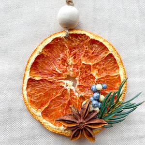 May include: A decorative dried orange slice, adorned with star anise, blue and white berries, and green sprigs. It has a wooden bead and twine for hanging. The orange slice measures approximately 8 cm in diameter.