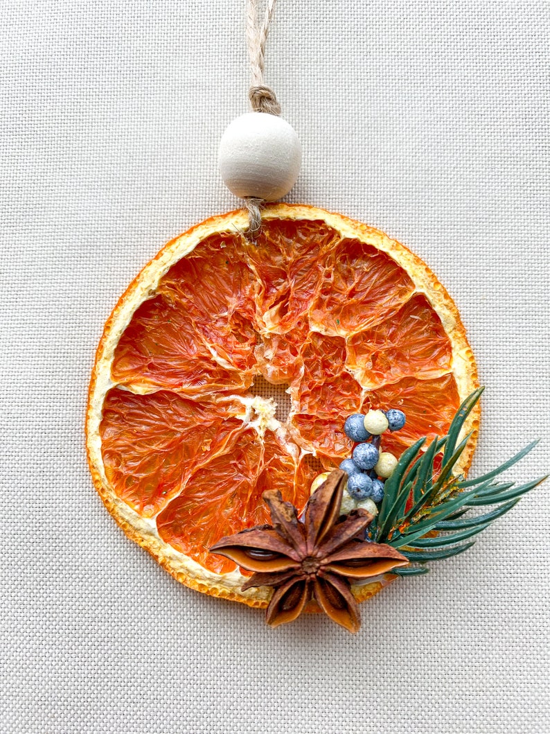 May include: A decorative dried orange slice, adorned with star anise, blue and white berries, and green sprigs. It has a wooden bead and twine for hanging. The orange slice measures approximately 8 cm in diameter.
