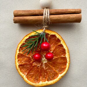 May include: A Christmas ornament made from a dried orange slice, decorated with sprigs of greenery and three red berries. Two cinnamon sticks are tied together with twine and a wooden bead. The ornament hangs from a natural twine string.