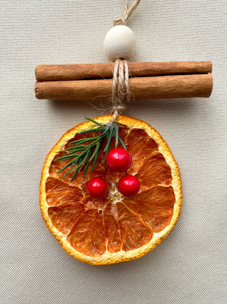 May include: A Christmas ornament made from a dried orange slice, decorated with sprigs of greenery and three red berries. Two cinnamon sticks are tied together with twine and a wooden bead. The ornament hangs from a natural twine string.
