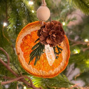 May include: A festive Christmas ornament made from a dried orange slice, a small pine cone, a clear crystal, and sprigs of greenery. It hangs from a twine string and a wooden bead. The background is a blurred Christmas tree with fairy lights.