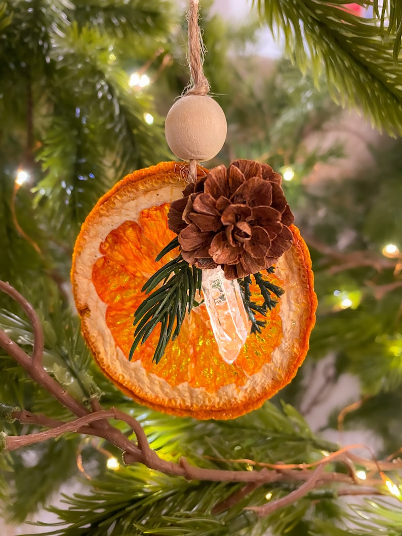 May include: A festive Christmas ornament made from a dried orange slice, a small pine cone, a clear crystal, and sprigs of greenery. It hangs from a twine string and a wooden bead. The background is a blurred Christmas tree with fairy lights.