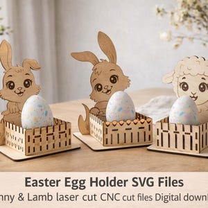 Puede incluir: Tres portahuevos de Pascua de madera con diseños de conejos y corderos. Cada soporte tiene una caja decorativa en forma de valla que contiene un huevo moteado. Los soportes están hechos de madera clara y están sobre una superficie de madera.