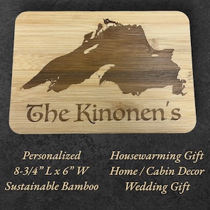 May include: A light brown wooden cutting board with a laser-engraved outline of the Upper Peninsula of Michigan. The text "The Kinonen's" is engraved below the outline. The board is 8-3/4 inches long and 6 inches wide. It is made from sustainable bamboo and is suitable for housewarming, home decor, or wedding gifts.