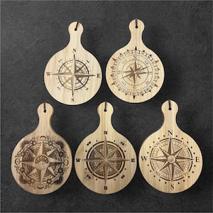 May include: Set of five wooden cutting boards with compass designs. Each board has a handle and a circular design with a compass rose and directional markings. The boards are made of light brown wood.