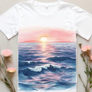 K&ouml;nnte beinhalten: Ein wei&szlig;es T-Shirt mit einem Aquarellbild eines Sonnenuntergangs &uuml;ber dem Meer. Das Bild zeigt einen rosa und orangenen Himmel, ein blaues Meer mit Wellen und eine strahlend gelbe Sonne.
