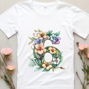 May include: White t-shirt with a floral design in shades of pink, yellow, blue, and green. The design features the number "6" surrounded by flowers.