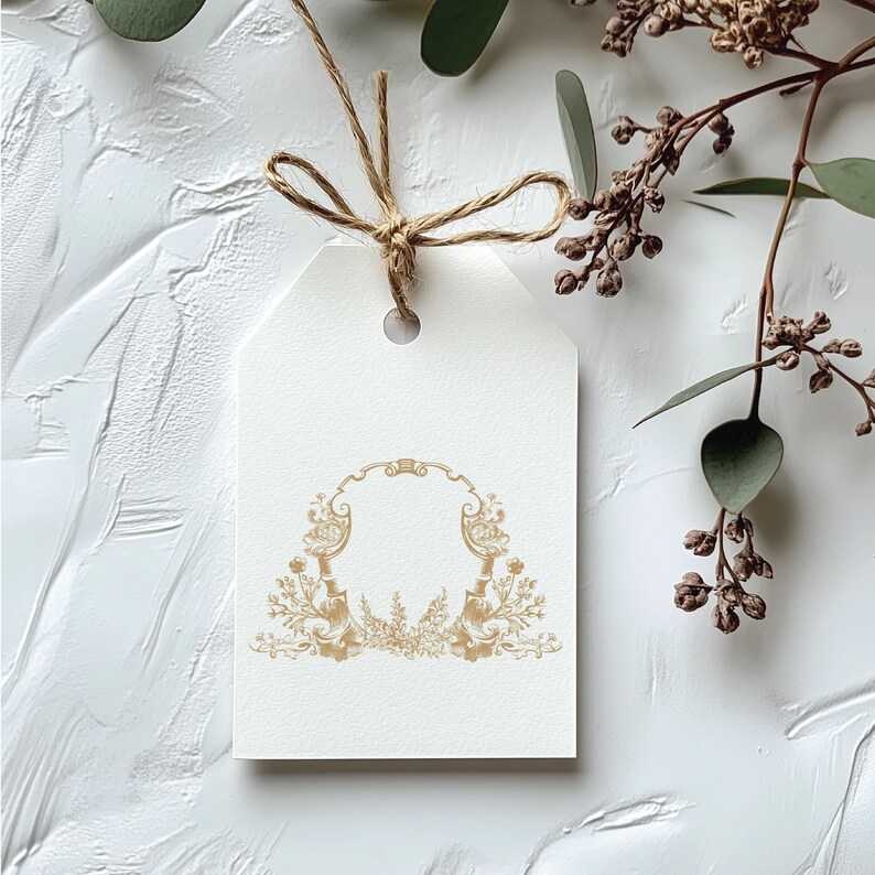 May include: A white gift tag with a gold ornate frame design. The frame is made of two symmetrical floral motifs with a central arch. The tag is tied with a brown twine bow.