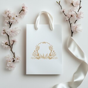 May include: White gift bag with a gold ornate frame design. The bag has a white satin ribbon tied around it.