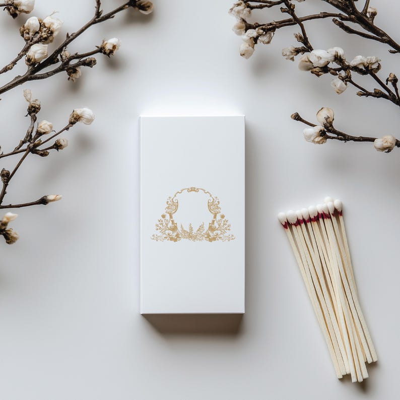 May include: A white box with a gold floral design on the front. The box is sitting on a white surface. There are white cotton branches on either side of the box. A bundle of wooden matches with red tips is on the right side of the box.